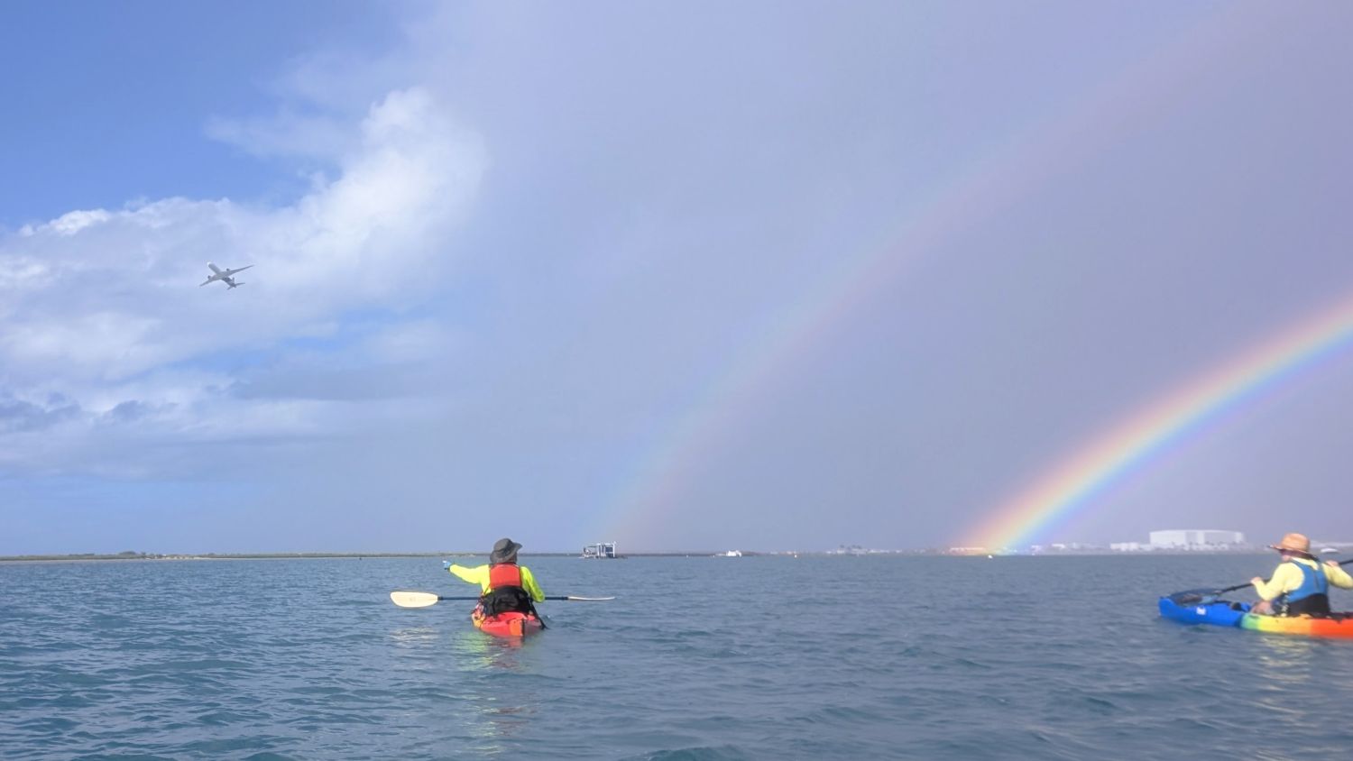 Two kayakers on flat water with a double rainbow and jet plane in the background